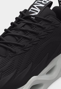 Μαύρο αθλητικό παπούτσι με υφασμένες πλευρές, παχύ λευκό πέλμα με ανοίγματα, μαύρα κορδόνια και μια ετικέτα με το λογότυπο στο γλώσσα.