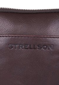 Pochette in pelle marrone scuro con logo "STRELLSON" in rilievo, texture liscia e chiusura a zip lungo il bordo superiore.