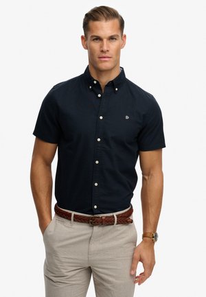 Hombre con camisa azul marino de manga corta abotonada, pantalones beige, cinturón marrón trenzado y reloj marrón, de pie con las manos en los bolsillos.