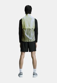 Letvægts, reflekterende, gul vest med sort bund, der har høj krave og baglomme, parret med sorte shorts og atletiske sko.