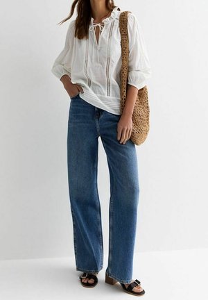 Frau trägt eine weiße, langärmelige Bluse mit Rüschen, weite blaue Jeans, schwarze Sandalen und trägt eine geflochtene braune Schultertasche.