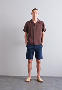 Braunes kurzärmliges Hemd mit einem Reverskragen, kombiniert mit navyblauen Shorts. Hellbraune Slippers mit einer strukturierten Oberseite und brauner Sohle.