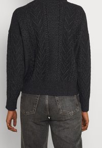 Pull noir en maille avec motifs torsadés texturés, col rond et coupe courte. Porté avec un jean taille haute gris foncé.