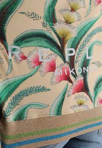 Blommig mönsterbärande väska i beige med gröna blad och rosa blommor. Har metalliskt guld och färgglada randiga detaljer i botten.