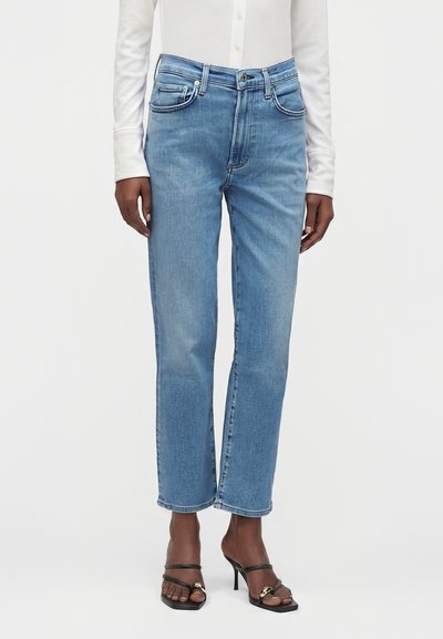 Person iført lyseblå straight-leg jeans, hvid langærmet skjorte og sorte højhælede sandaler med stropper, står mod en ensfarvet baggrund.