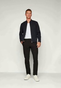 Veste de bombardier navy avec fermeture éclair argentée, t-shirt à col rond blanc, pantalon noir slim-fit et baskets blanches. Tissu lisse, coupe moderne.