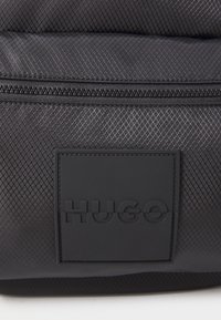 Primo piano di una pochette in tessuto nero testurizzato con zip e una toppa quadrata nera impresso con la parola "HUGO" sul davanti.