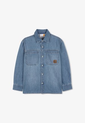Chemise en denim bleu clair à manches longues, avec deux poches poitrine, fermeture boutonnée sur le devant, et un patch logo marron sur la poche gauche.