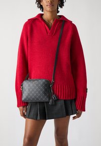 Maglione rosso a maglia con scollo a V e orlo a coste, abbinato a una borsa a tracolla nera con motivo, dotata di una frangia e accessori in oro.
