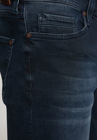 Jeans din denim albastru închis, cu o croială ajustată, având un efect subtil de uzură, un buzunar pentru monede și o cusătură contrastantă. Accesoriile includ nituri din cupru.