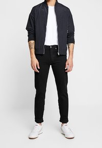 Chaqueta bomber negra, camiseta blanca, jeans negros y zapatillas blancas. La ropa tiene un corte casual con cierre frontal y puños enrollados.