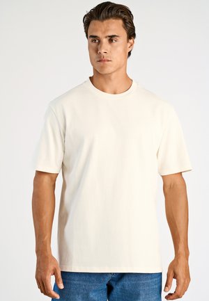 Young man with dark hair wearing a plain off-white short-sleeve T-shirt and blue jeans, standing against a plain light background.