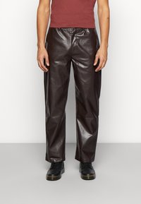 Pantalones de cuero marrón con un acabado brillante, que presentan un corte relajado y una cintura tradicional, combinados con zapatos negros.