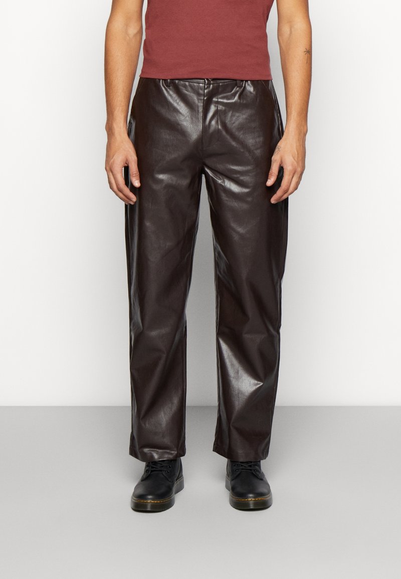 Pantalones de cuero marrón con un acabado brillante, que presentan un corte relajado y una cintura tradicional, combinados con zapatos negros.
