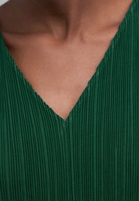 Närbild av en persons nacke och övre bröst som bär ett mörkgrönt veckat V-ringat plagg.