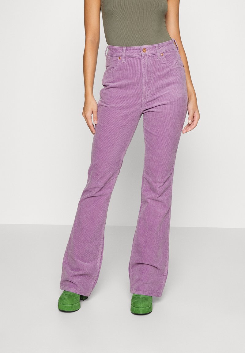 Personne portant un pantalon en velours côtelé lavande taille haute, un débardeur vert olive et des chaussures compensées en daim vertes, debout sur un sol gris.