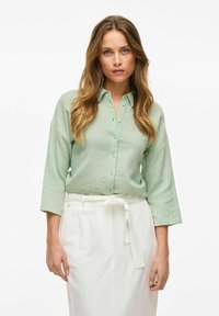 Camicia button-up verde chiaro con maniche a tre quarti, realizzata in tessuto testurizzato, abbinata a una gonna bianca con vita annodata.