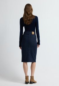 Tummansinisestä denimitikusta valmistettu kynähame, jossa on takataskut ja vyötäröllä ruskea nahkalappu, yhdistetty pitkähihainen laivastonsininen paita ja beiget nilkkurit.