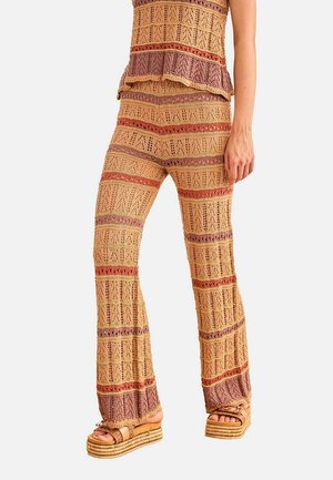 Mujer viste pantalones de punto a crochet con rayas en beige, marrón y óxido, y top sin mangas a juego, con sandalias de plataforma sobre fondo blanco.