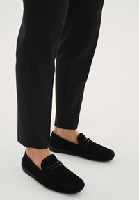 Mustat mokkanahkaiset loafersit, joissa on litteä pohja, sileä pinta ja koristeellinen remmi kengän päällä, yhdistetty räätälöityihin mustiin housuihin.