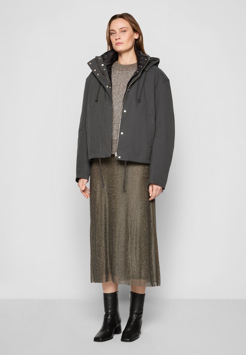 Graue, verkürzte Jacke mit Kapuze, Kordelzug und Druckknöpfen; über einem metallic gestrickten Pullover und einem schimmernden, plissierten Rock getragen; kombiniert mit schwarzen Stiefeletten.
