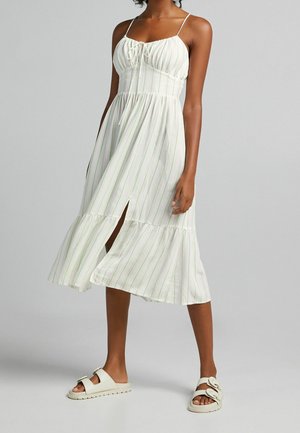 Vrouw draagt een witte mouwloze midijurk met dunne groene strepen en witte instapsandalen met gespen.