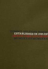 Olivengrønt stof med ribbet struktur og trykt tekst i marineblå, rød og hvid, der detaljerer "Tommy Hilfiger Established 1985."