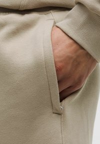 Beige mikina s blízkym pohľadom na látku a rukou v vrecku. Viditeľný textúrovaný povrch a detail rebrového manžety blízko zápästia.