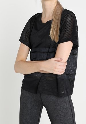 Žena v černém síťovaném tričku s krátkými rukávy a tmavě šedých legínách, s překříženýma rukama, na jednobarevném světlém pozadí.