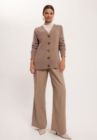 Khaki kardigan z dekoltem w kształcie litery V, pięcioma dużymi guzikami, ściągaczami przy mankietach i u dołu. Stylizowany z szerokimi beżowymi spodniami i spiczastymi beżowymi szpilkami.