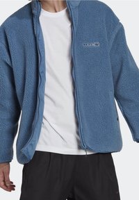 Ljusblå fleecejacka med texturerad yta, hel dragkedja, två sidofickor och en liten logotyp på bröstet.