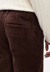 Brune kordfløjspants med en tekstureret overflade, der har en baglomme og et logo-tag. Livremmen har bælteløkker for ekstra støtte.