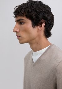 Beige V-hals trui gemaakt van zachte gebreide stof, met een subtiele textuur en gestikte details, gedragen over een wit shirt met lange mouwen.