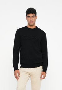 Maglione nero lavorato a maglia con collo rotondo e logo "GUESS" in rilievo sulla parte anteriore. Presenta polsini e orlo a coste per un look aderente.