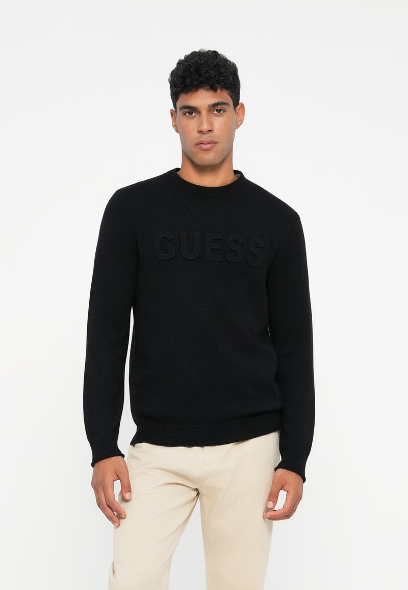 Maglione nero lavorato a maglia con collo rotondo e logo "GUESS" in rilievo sulla parte anteriore. Presenta polsini e orlo a coste per un look aderente.