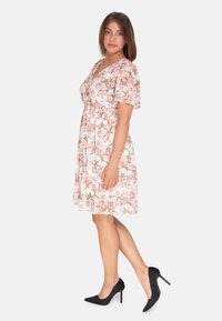 Blumenkleid mit kurzen Puffärmeln, taillierter Taille und knie-langer Saum. Rosa Blumen auf weißem Hintergrund. Kombiniert mit schwarzen Stilettos.