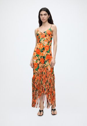 Femme aux longs cheveux foncés portant une robe orange à fleurs avec des bretelles spaghetti et un ourlet à franges, debout devant un fond uni.