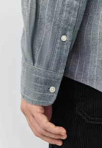 Cămașă cu dungi gri și albă dintr-un material texturat, având manșete cu butoane. O mână se odihnește pe o pereche de pantaloni din catifea în culoare neagră.
