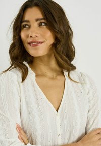 Blusa blanca con escote en V, tejido texturizado con patrones de diamantes y cierre de botones. Mangas largas con ligeros volantes en el escote.