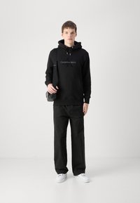 Zwarte hoodie met logo-opdruk, gecombineerd met losse zwarte broek en witte sneakers. Model draagt een kleine zwarte schoudertas.