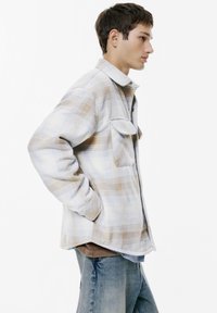 Checkered flannel shirt in light blue and beige tones, featuring a collar, front pockets, and a relaxed fit. Worn over a brown top.
