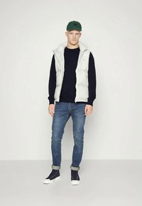 Joven vestido con una gorra verde, un chaleco acolchado blanco, una camiseta de manga larga negra, jeans azules arremangados y zapatillas altas negras, de pie contra un fondo liso.