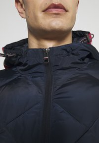 Man i en navyblå fodrad jacka med dragkedja och huva, där snoddarna är synliga vid halsen.