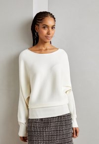 Pull blanc côtelé avec un large col et des manches longues légèrement amples, associé à une jupe texturée noir et blanc.