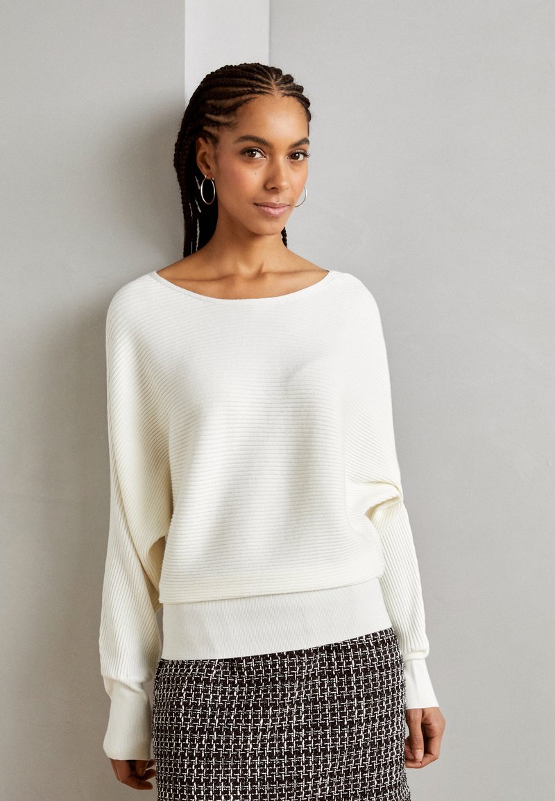 Pull blanc côtelé avec un large col et des manches longues légèrement amples, associé à une jupe texturée noir et blanc.