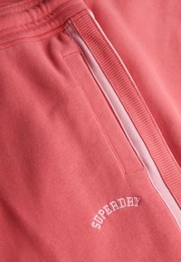 Primo piano di un tessuto corallo rosa con vita a coste, tasca laterale e testo ricamato bianco "SUPERDRY" curvato sopra la tasca.