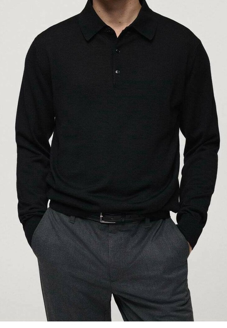 Homme portant un polo noir à manches longues rentré dans un pantalon gris avec une ceinture noire, mains dans les poches.