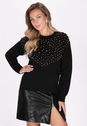 Donna con capelli lunghi e mossi che indossa un maglione nero con perline colorate e una gonna nera strutturata con una zip frontale e uno spacco.