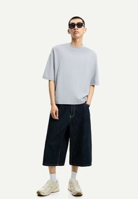 Siv t-shirt v prostem kroju, temno modri široki denim kratki hlače, bele nogavice in bež športni čevlji z rumenimi detajli. Črne sončne glasses.