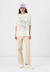 T-shirt surdimensionné beige clair avec des graphismes de palmier, associé à un pantalon en velours côtelé beige clair et des baskets blanches. Casquette lavande portée au-dessus.